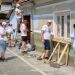 Group of volunteers painting the exterior wall of a building on a sunny day; one person on a ladder applying paint while others work nearby.