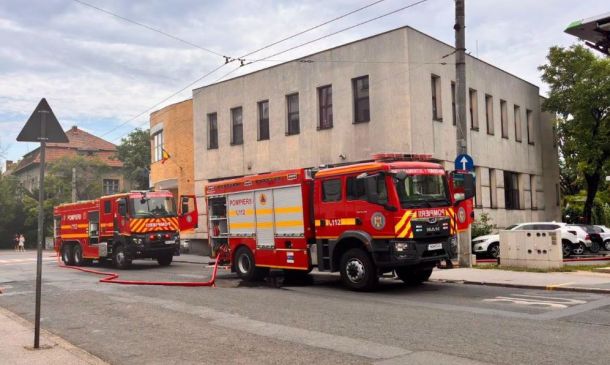 Two red fire trucks parked on a city street with hoses laid out near a beige multi‑story building under a cloudy sky, daytime scene.
