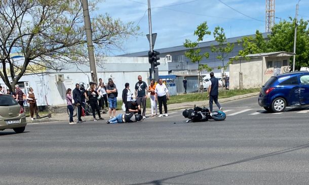 Crowd gathered at an intersection after a motorcycle crash; a rider lies on the pavement near a fallen bike while another motorcycle sits nearby and cars pass by.