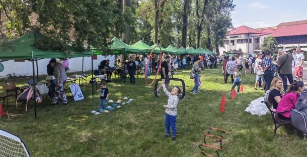 Outdoor community fair in a park with green vendor tents, families and kids playing games and chatting.