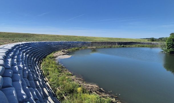 Curved wall of stacked concrete bags along a calm water body under a clear blue sky and grassy embankment beyond.