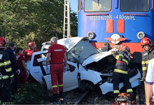 White car severely damaged after collision with a blue train, emergency responders in red and yellow gear assisting on the tracks.
