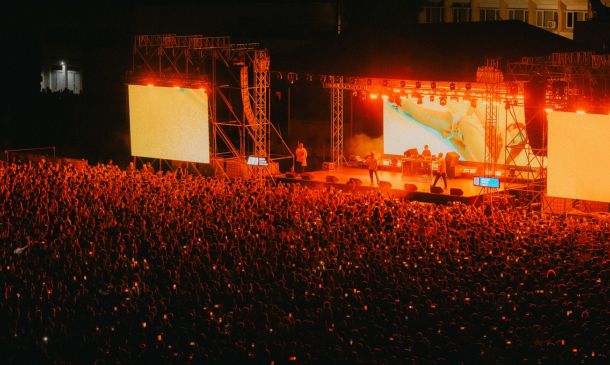 Outdoor night concert with a dense crowd facing a brightly lit stage and two large screens.