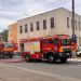 Two red fire trucks parked on a city street with hoses laid out near a beige multi‑story building under a cloudy sky, daytime scene.