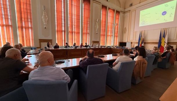 Formal meeting in a council chamber with people seated around a U-shaped wooden table and a large projector screen at the front.