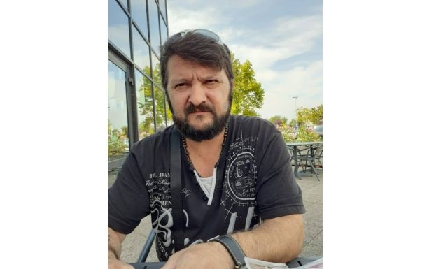 Bearded man wearing a black graphic shirt sits at an outdoor cafe, sunglasses on his head.