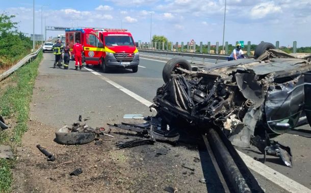 Emergency responders and a red ambulance on a highway beside a severely damaged overturned car and debris on the road flank a crash scene.