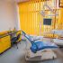 Modern dental treatment room with a blue reclining chair, yellow blinds, and dental equipment nearby.