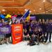 Group of students in purple team shirts posing with banners and a Ukrainian flag at a robotics competition, trophies on the floor, indoor arena backdrop