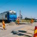 Blue locomotive at a rural railroad crossing under construction, with orange-yellow striped barriers and warning signs nearby.