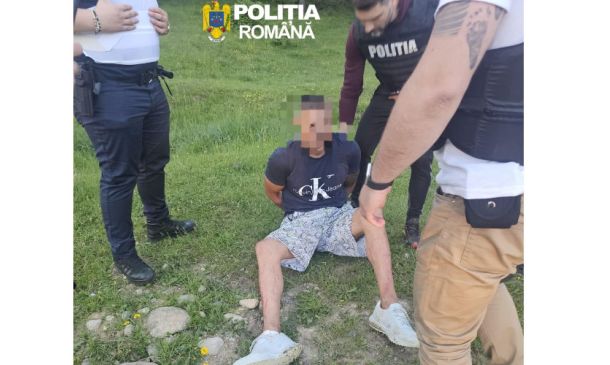 A man sits on grass with hands behind his back as three Romanian police officers surround him; face blurred, police logos visible above.