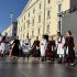 Group of people in traditional folk costumes performing a dance on an outdoor stage in front of a yellow historic building.