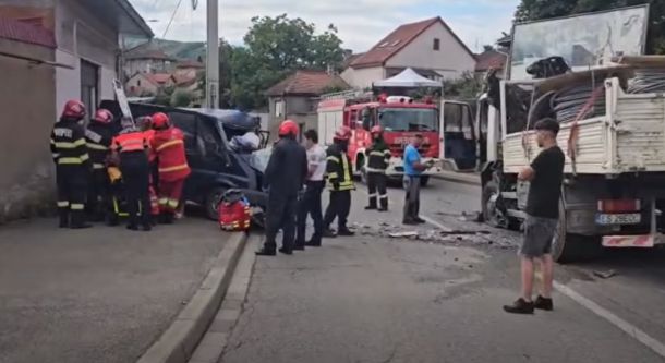 Firefighters and responders work at a car crash on a street with a tow truck and debris nearby, a fire truck in the background.