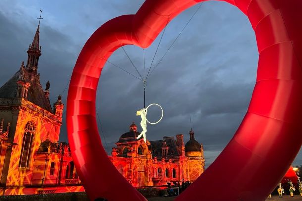 Acrobat on a hoop suspended by cables inside a large red inflatable ring, with a warmly lit historic building in the background at dusk.