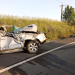 Wrecked white sedan after a severe crash on a road with emergency responders tending to a person nearby on the grass verge to the right of the scene.