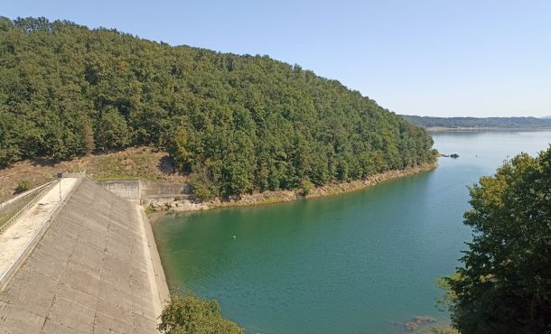 Reservoir scene: a sloped concrete dam on the left, calm turquoise water, and a dense forested hill rising behind.