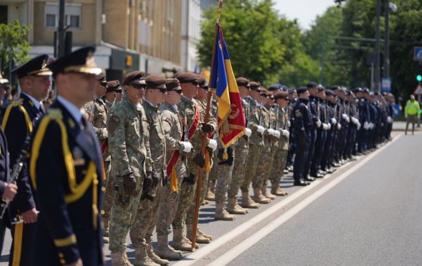 Line of soldiers in uniform standing at attention during a parade, one holding a blue-yellow-red flag.