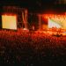 Outdoor night concert with a dense crowd facing a brightly lit stage and two large screens.