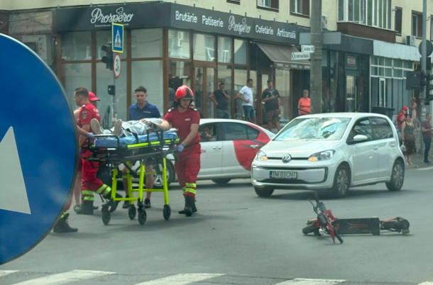 Paramedics in red uniforms wheeling a patient on a stretcher across a city street as a red motorcycle lies on its side nearby, with bystanders and storefronts in the background.