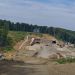 Construction site on a hillside with a tunnel entrance under an embankment, dirt roads and equipment nearby