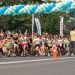 Children start a road race under a blue‑green balloon arch; spectators and organizers watch nearby.