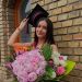 Graduate woman wearing a cap and gown holding a large bouquet of pink and white flowers outside a brick building.