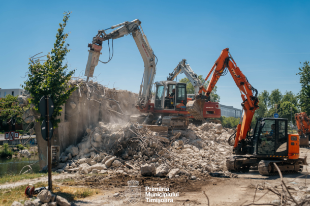 Two excavators demolish a concrete wall at a sunny construction site, dust rising over a pile of rubble; Primăria Municipiului Timișoara logo visible at the bottom.