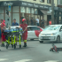 Paramedics in red uniforms wheeling a patient on a stretcher across a city street as a red motorcycle lies on its side nearby, with bystanders and storefronts in the background.