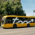 Yellow city bus with a large white logo on its side parked along a street with trees and buildings in the background.