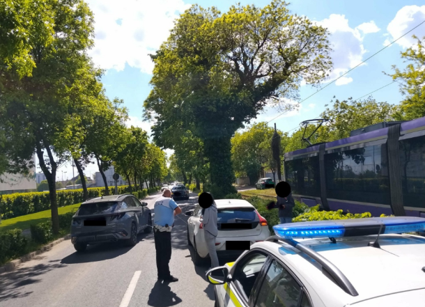 Police officer talks to the driver of a white car on a sunny tree-lined street; a tram is on the right and police car lights are visible in the foreground.