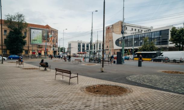 Public square with wooden benches, a dirt circular planting bed, and a street with buses and pedestrians.
