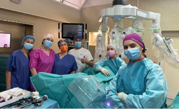 Surgical team in masks and gowns pose in an operating room beside a robotic surgery system with multiple robotic arms.