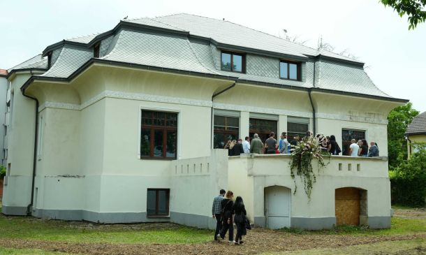 Group of people gathered on a balcony in front of a cream-colored house with a gray tiled roof and decorative molding.