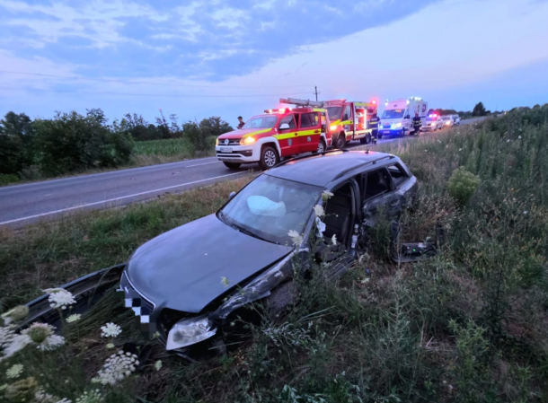 Gray sedan crashed off a rural road into tall weeds, front end damaged, with emergency vehicles and responders arriving in the background at dusk.