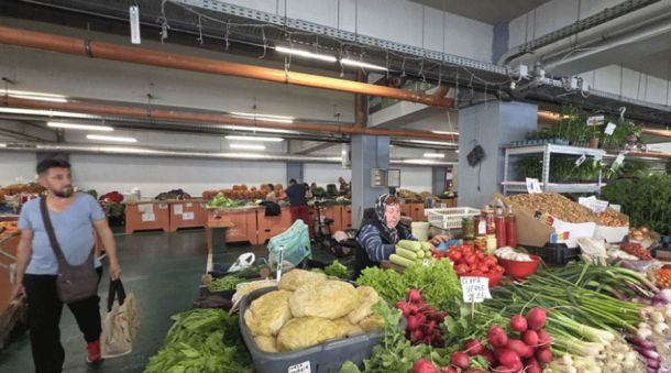 Indoor market stall with fresh vegetables displayed in crates; a vendor wearing headphones sits behind the produce while shoppers browse in the background.
