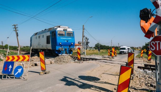 Blue locomotive at a rural railroad crossing under construction, with orange-yellow striped barriers and warning signs nearby.