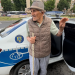 Older man with a cane and hat stands by the open rear door of a white and blue police car on a city street.