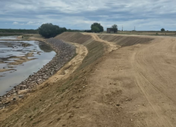 Dirt road along a sloped, rocky riverbank with calm water to the left and distant trees under a cloudy sky.
