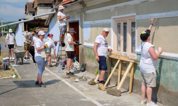 Group of volunteers painting the exterior wall of a building on a sunny day; one person on a ladder applying paint while others work nearby.