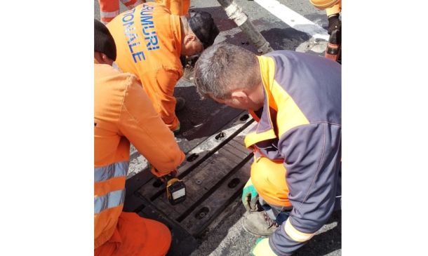Construction workers in high-visibility orange gear operating a handheld device over an open road trench area.