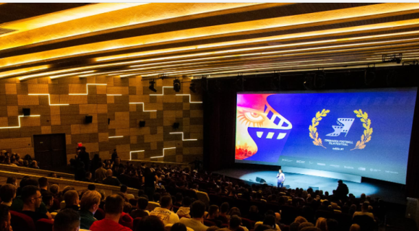 A crowded theater auditorium with attendees looking toward a brightly lit stage and a large colorful screen displaying festival graphics on the right side.