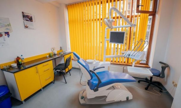 Modern dental treatment room with a blue reclining chair, yellow blinds, and dental equipment nearby.