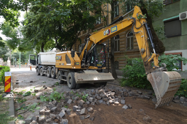 Yellow excavator and dump truck parked on a city street as bricks and dirt are excavated nearby