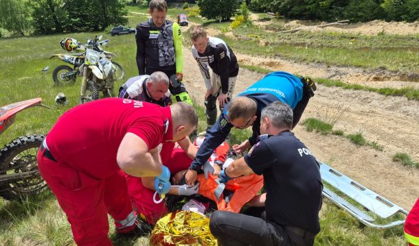 Emergency responders in red and dark uniforms attend to a person on the ground in a grassy off-road area, with dirt bikes nearby.