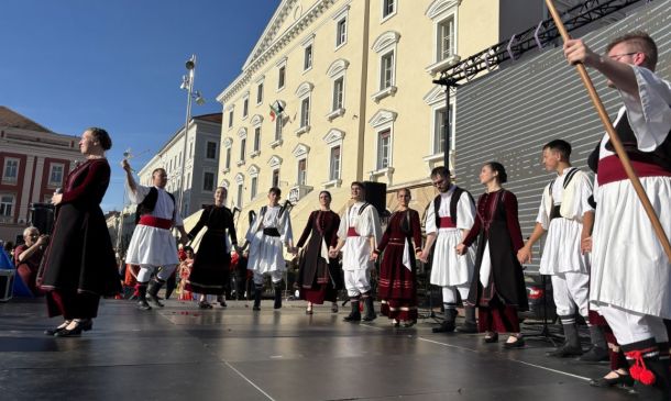 Group of people in traditional folk costumes performing a dance on an outdoor stage in front of a yellow historic building.