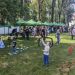 Outdoor community fair in a park with green vendor tents, families and kids playing games and chatting.
