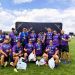 Rugby team in purple jerseys posing on a sunny field with medals and a trophy after winning.