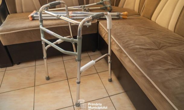 Folding aluminum walker with rubber tips set up in a tiled living room beside a beige sectional sofa.