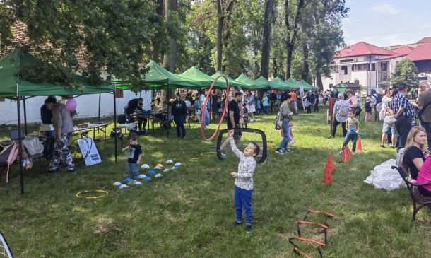 Outdoor community fair in a park with green vendor tents, families and kids playing games and chatting.