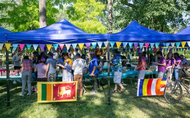 Outdoor park festival under blue canopies with colorful pennant bunting and people browsing vendor tables; Sri Lanka flag visible at front fence.